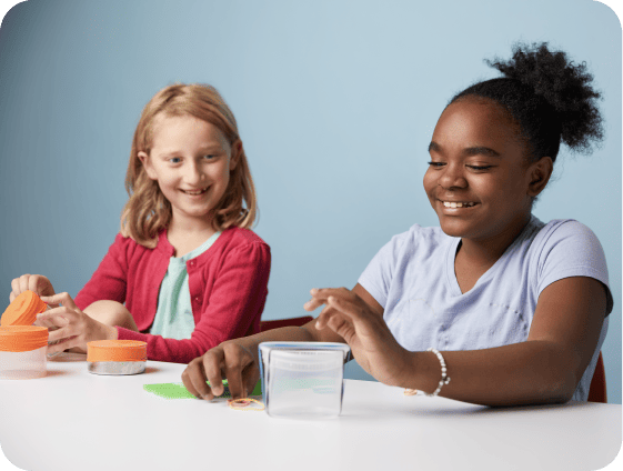 Two girls sit at a table, immersed in an elementary school science curriculum activity. One is handling a container while the other smiles, playing with a curious green object. The light blue background complements their lively learning experience.